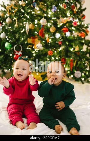 Baby twin siblings together laughing in front of christmas tree Stock Photo