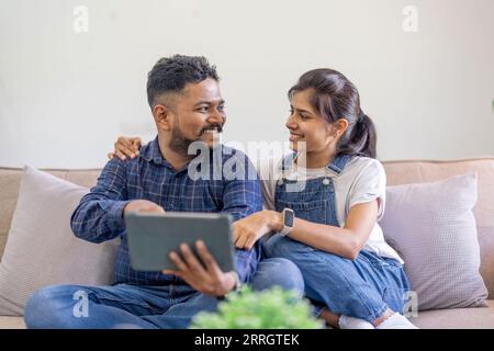 Beautiful indian spouses rest on sofa at home, using digital tablet explore the world of social media and shopping online together Stock Photo