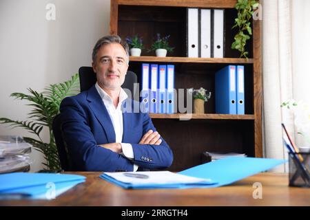 Smiling entrepreneurial businessman in his home office wearing blue suit and documents on wooden table and bookshelves background. Stock Photo