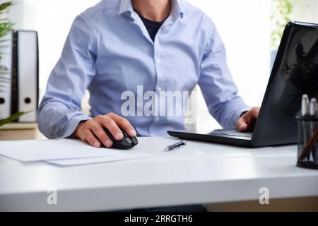 Detail of a man working with a laptop sitting in an office chair using the mouse on a white work table Stock Photo