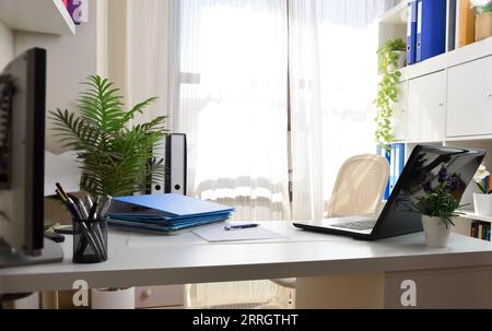Home office with white furniture with folders on table and shelves and laptop without people. Stock Photo