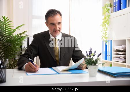 Smiling man in gray suit, white shirt and gold tie writing on a document in a bright office with white furniture. Stock Photo