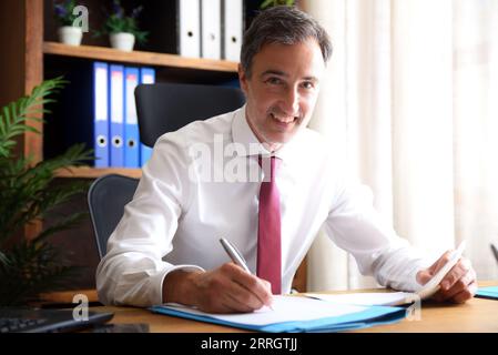 Smiling man in white shirt and red tie writing on a document and looking working in an office with wooden furniture Stock Photo
