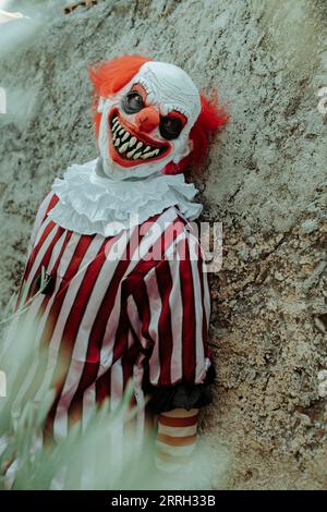 a mad evil redhead clown, wearing a white and red striped costume, and leaning on the rustic wall of an old house outdoors, stares at the observer wit Stock Photo