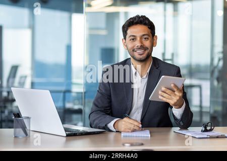 Portrait of young businessman with tablet computer inside office, hispanic man smiling and looking at camera, working with laptop, using app on tablet. Stock Photo