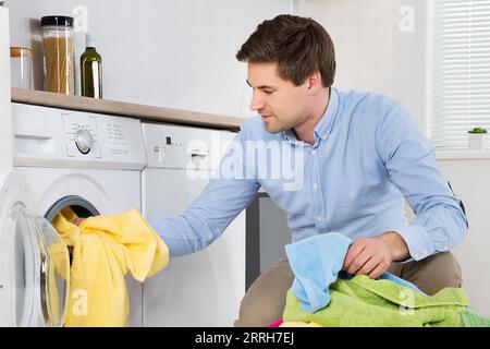Young Man Loading Clothes Into Washing Machine In Kitchen Stock Photo
