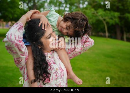 Happy young indian mother carrying child daughter on her shoulders at summer park.Family and motherhood concept. Stock Photo