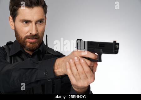 Focused bearded officer taking aim, by holding handgun both hands in studio. Portrait of caucasian cop keeping gun, zeroing in side, looking away, on gray background. Concept of danger work, weapon. Stock Photo