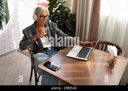 cute european gray-haired senior business woman sits in a cafe and watches a video on a laptop using headphones Stock Photo