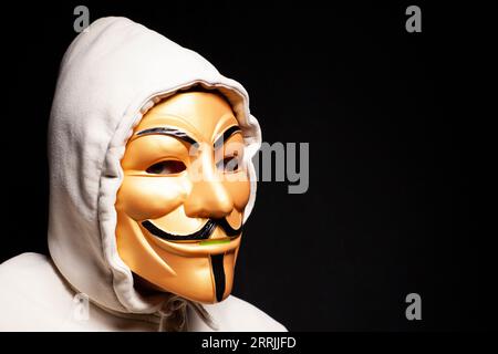 Moscow, Russia, September 03, 2023: Hacker wearing hood and gold anonymous mask on black background. Stock Photo