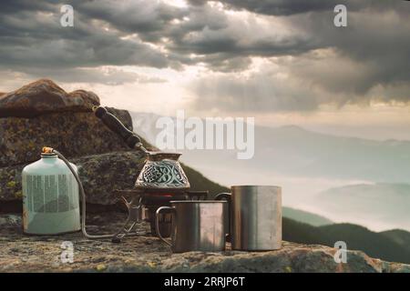 Hot outdoor coffee with a view. Camping stove with boiling coffee cezve and stainless travel mugs on the stone on a mountain top. Stock Photo