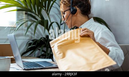 Employee female working at the desk talking with clients using headset and microphone and laptop. Customer care service business. One adult woman work online and prepare package for shipment remote Stock Photo