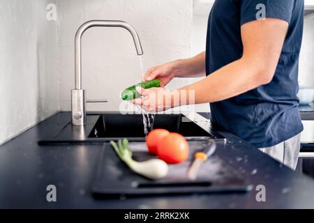 Washing vegetables in kitchen sink with water. Food preparation. Man cooking. Making salad. Cucumber in hand under faucet tap. Vegan or vegetarian. Stock Photo