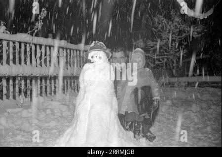 England. c.1960 – A young boy and his father posing next to a snowman they have built in their garden during a snow flurry. A trilby hat and a pipe have been added to the snowman as embellishment. The boy is wearing a waterproof coat and a knitted woollen hat. Stock Photo