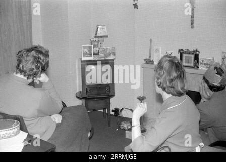 England. c.1960 – A family are sitting in a lounge watching television on Christmas Day. The wooden-cased television is a TV43 model manufactured by Bush during the 1950s. The man is wearing a tissue paper hat from a Christmas cracker. On top of the television is a calendar displaying the date of 25th December.  The room has a tiled fireplace surround which is decorated with greetings cards, candles and holly. Stock Photo