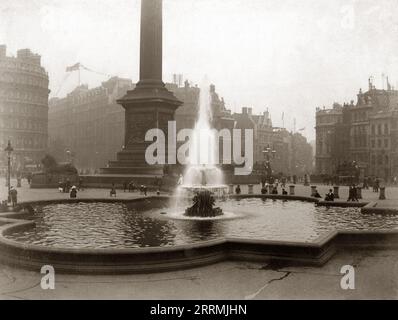 London, England. 1911 - A view of one of the fountains and the base of Nelson’s Column at Trafalgar Square, London. The photograph shows the original fountain centrepiece dating from the 1840s. This was replaced following the Second World War with a new design by Sir Edwin Lutyens. A group of children are gathered around the fountain.  Horse-drawn omnibuses can be seen passing by. The roof of a building in The Strand, visible in the distance is decorated with a giant White Ensign flag and bunting in preparation for the coronation of King George V in June 1911. Stock Photo