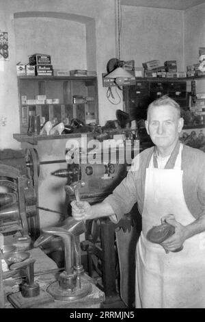 England. c.1960 – A cobbler posing for a photograph in his workshop. He is wearing an apron whilst holding a cobbler’s hammer and a boot. He is standing in front of a bench, mounted on which is a selection of steel shoe lasts. In the background are specialist machinery and shelves containing shoes and a variety of supplies. Stock Photo