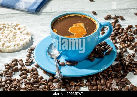 Morning coffee. A blue cup of coffee with foam on a wooden table surrounded by scattered coffee beans. In the background there is a rice cake and a co Stock Photo