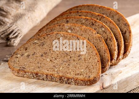 Slices of fresh rye bread with grains lie on the kitchen board, in the background there are ears of barley and a burlap napkin, close-up rustic style. Stock Photo