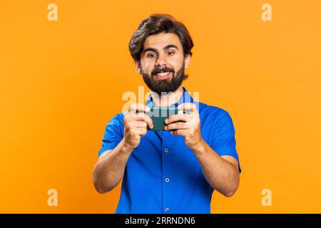Worried funny addicted man enthusiastically playing racing video games on mobile phone. Happy middle eastern guy using smartphone gadget app with drive simulator isolated on orange studio background Stock Photo
