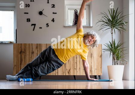 Active senior man in yellow tshirt taking care of his health by exercising and working out. Doing a side plank position for strong core on a mat in do Stock Photo