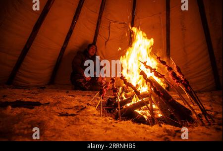 230113 -- GENHE, Jan. 13, 2023 -- Dalinma, Juele s cousin, cooks roasted meat for dinner in a traditional Ewenki tent at a temporary herding station at the Jinhe forest range near Genhe City, north China s Inner Mongolia Autonomous Region, Jan. 11, 2023. Every two or three days, Juele Bulituotian will head for the Jinhe forest range about 80 kilometers from Genhe City. That s where the 39-year-old Aoluguya Ewenki herder, wearing a traditional fur coat, will find his foraging reindeer. Instead of keeping reindeer in a barn, Ewenki herders let the animals live in the forest and check on them eve Stock Photo