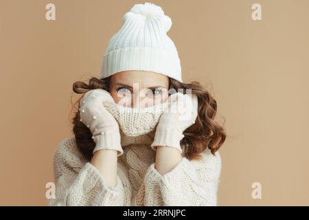 Hello winter. elegant middle aged woman in beige sweater, mittens and hat isolated on beige background wrapped in a collar. Stock Photo