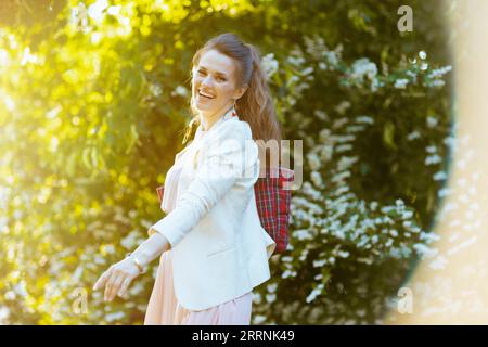 smiling elegant 40 years old woman in pink dress and white jacket in the city against greenery. Stock Photo