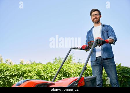 Smiling male gardener in protective glasses trimming turf with electric mower in sunny day. Low angle view of bearded guy using lawn trimmer, working in garden, against blue sky. Concept of work. Stock Photo