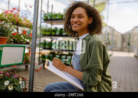 Woman florist writing down plant inventory on clipboard in flower garden center Stock Photo