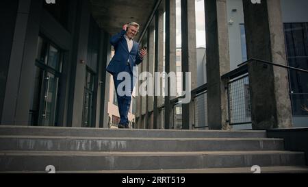 Old businessman middle-aged Caucasian employee senior business man worker listening to music in headphones holding mobile phone dancing walking down Stock Photo