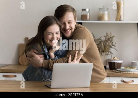 Positive overjoyed young husband and wife hugging at laptop Stock Photo
