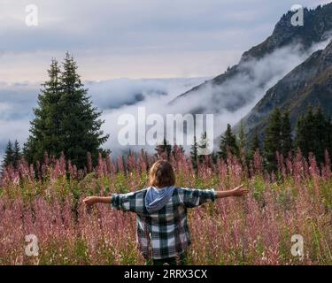 Happy female tourist in the mountains enjoys nature, against backdrop of mountains, fog and forest, with her back to the camera and arms outstretched Stock Photo