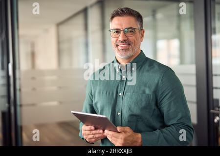 Older smiling businessman holding tablet standing in office. Portrait. Stock Photo