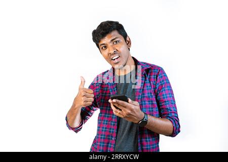 A young man in his 20s is holding a mobile phone in his right hand and giving a thumbs up with his left hand. He is smiling and looking at the phone s Stock Photo