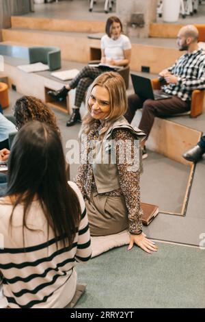 Two female employees having a conversation. Blonde, middle-aged woman hearing pleasant news by her brunette coworker Stock Photo