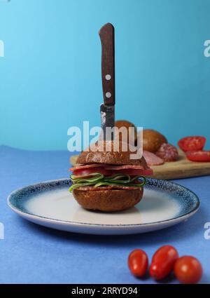 a burger sandwich with sausage vegetables cucumber tomato with buckwheat rye bun lies on a blue plate on a blue background. Vertical photo. cherry tom Stock Photo