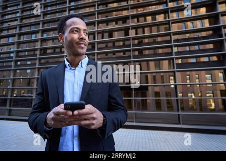 Young Latin professional male looking curious to side using smart phone center building background Stock Photo