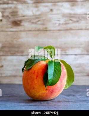 A single peach with leaves rests on a slate table with a rustic wood plank background Stock Photo