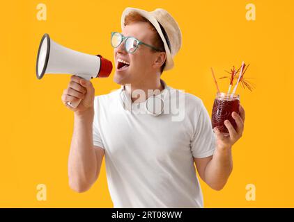 Young man with glass of juice shouting into megaphone on yellow background Stock Photo