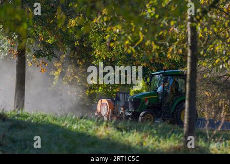 Small tractor of the municipality with front mounted leaf blower device driving through public park cleaning the walkways on sunny autumn day in Stock Photo