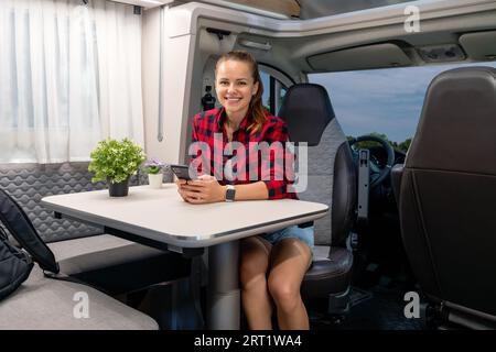Young adult brunette woman wearing plaid shirt sitting inside the motor home while parked in a camper van parking place. RV rental. Stock Photo