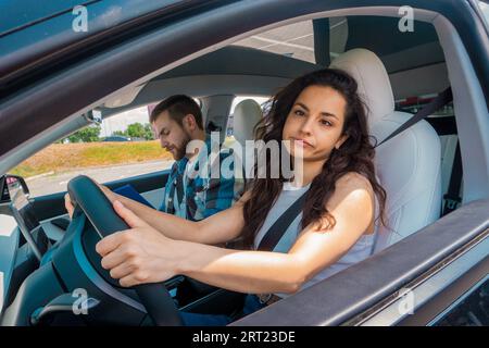 Young woman with a funny face sitting in the car and holding the steering wheel with her hands, instructor making notes. Driving test, driver courses Stock Photo