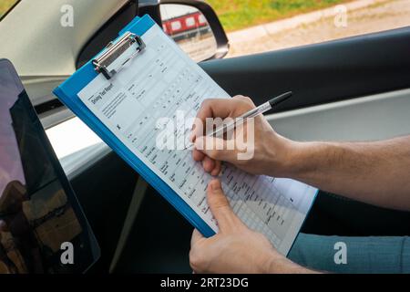 Male instructor filling out the driving test report while sitting in the car during driving exam. Close up. Driving test, driver courses, exam concept Stock Photo