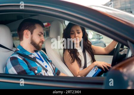 Male instructor writing in the form after drive exam, young woman worrying, sitting on the drive seat. Driving test, driver courses, exam concept Stock Photo