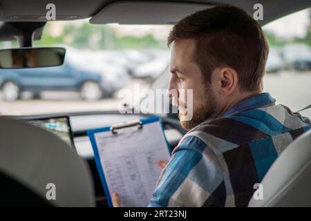 Instructor holding drive test report while sitting on the passenger seat. Back view. Driving test, driver courses, exam concept Stock Photo