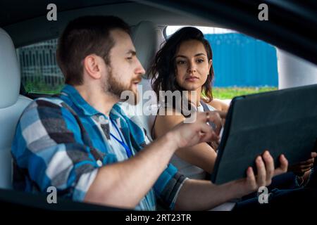 Young male instructor holding digital tablet and explaining female student sitting in the car during driving lesson. Driving test, driver courses Stock Photo