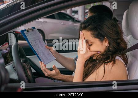 Tired young woman sitting in car covering face in hands, instructor sitting on the passenger seat and holding test report. Driving test, driver Stock Photo