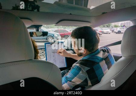 Back view of male instructor sitting on the passenger seat while holding drive test report, showing to lady. Driving test, driver courses, exam Stock Photo