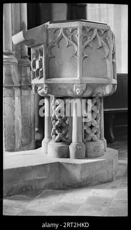 St Andrew's Church, Churchyard, Market Place, Castle Combe, Wiltshire, 1940-1949. Detail of a 14th century octagonal stone font with panelled reliefs on the bowl and openwork patterns on the pillars. On the left side of the bowl is a book rest above a panel with a floral design. The other panels on the bowl have reliefs of Gothic archways, and there are small figures on the underside of the bowl at each corner. The plinth is octagonal, with the side under the book rest extended for reading. Stock Photo